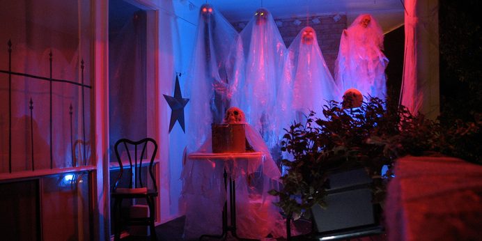 The original balcony haunt in 2007: white gauze ghosts with glowing eyes hung in blue and red light on a condo balcony, with a wrought-iron rail, a skull on a draped table, and potted plants.